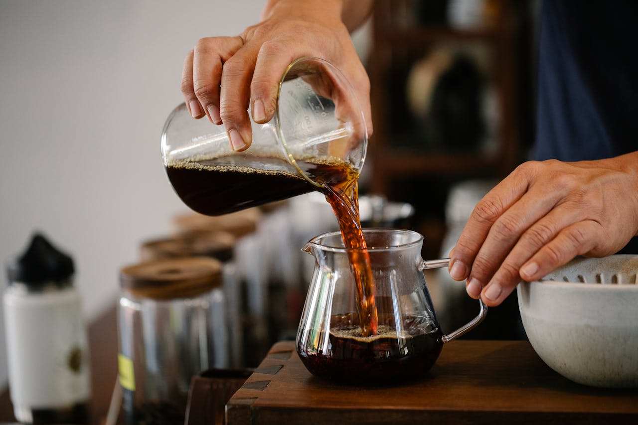 Crop faceless male worker pouring aromatic hot coffee from glass measuring pitcher into pot placed on wooden counter in cafe